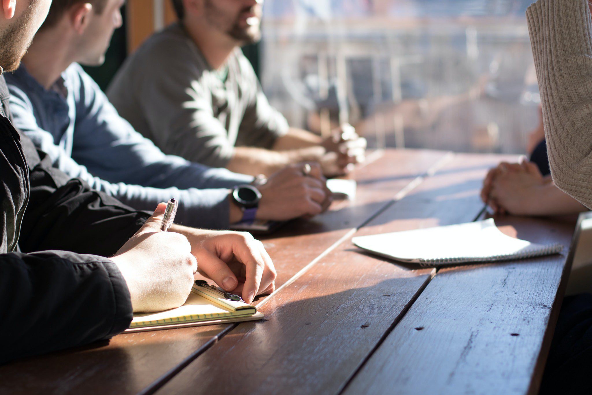 Four people sit around a wooden table with notebooks, engaged in a meeting or discussion, with sunlight streaming in through a window.