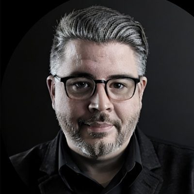 A man with gray hair, glasses, and a beard wears a black shirt and blazer, posing against a dark background—a portrait reminiscent of insightful Case Stories.