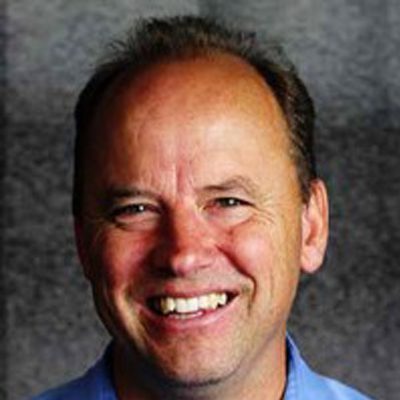A smiling middle-aged man with short hair wearing a light blue shirt, posed in front of a dark, textured background, ready to share his inspiring Case Stories.