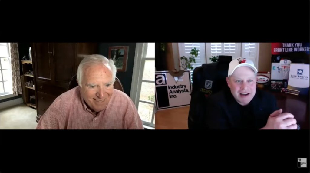 Two men, Ed McLaughlin and Shawn Cashmark, are on a video call from their respective home offices. One wears a pink shirt and smiles, while the other—donning a black jacket and white cap—sits in front of signs reading Thank You Front Line Workers, Industry Analysts, Inc., and EKM Global.