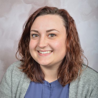 A woman with wavy, shoulder-length brown hair smiles at the camera. She is wearing a gray cardigan over a blue top, and is posed in front of a light, textured background—capturing the warmth and authenticity often found in our Case Stories.