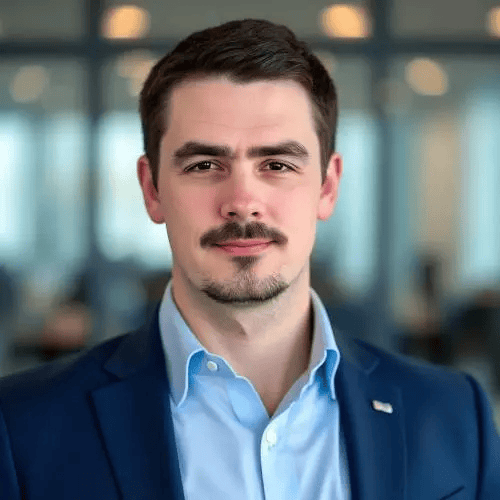 A man with short brown hair and a mustache wearing a blue suit jacket and light blue shirt stands in an office with blurred background.