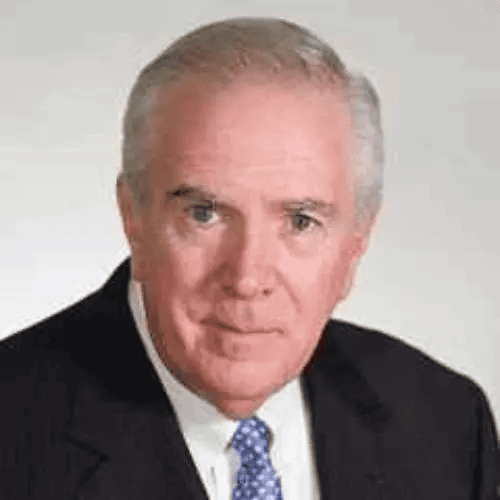 An older man with short gray hair wearing a black suit, white shirt, and blue patterned tie, looking directly at the camera against a plain light background.