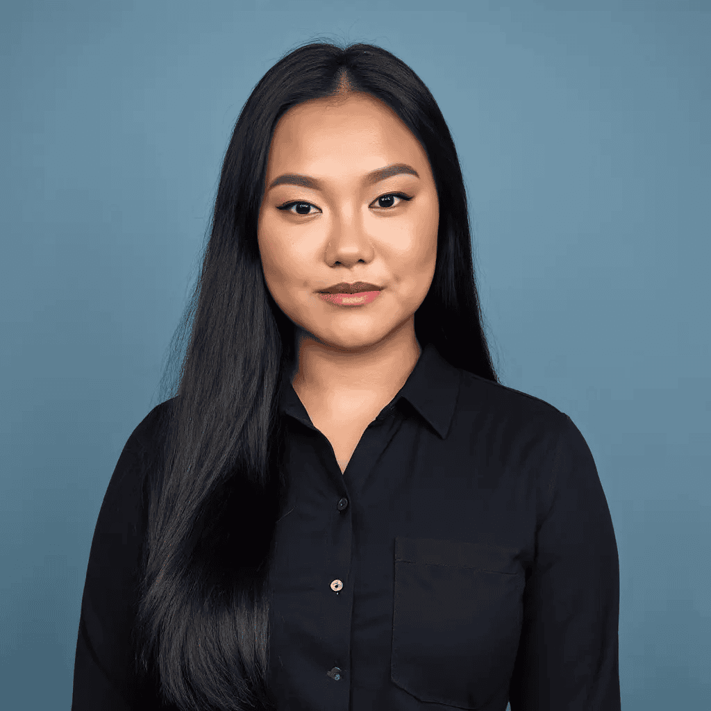 A woman with long black hair and a black button-up shirt stands in front of a plain blue background, looking directly at the camera with a neutral expression.