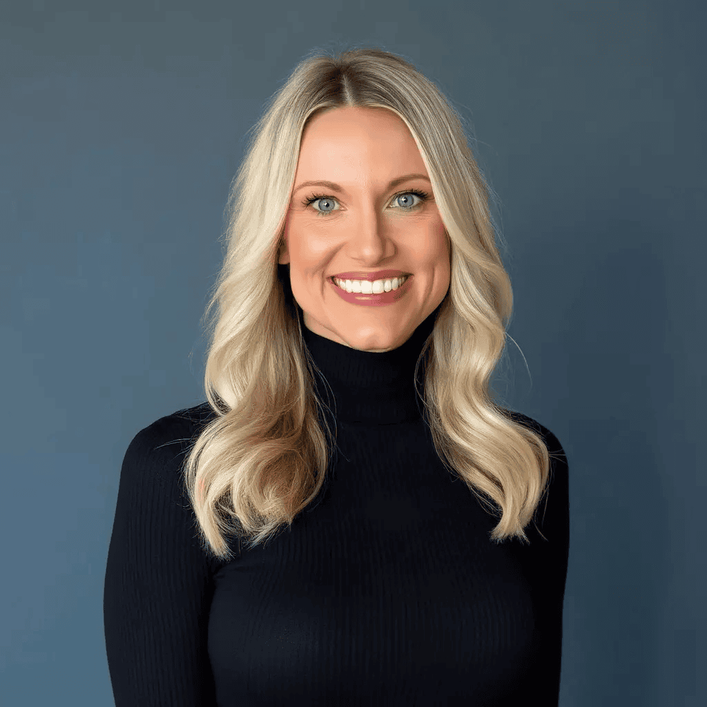 A smiling woman with long blonde hair wearing a black turtleneck sweater stands in front of a plain gray background.