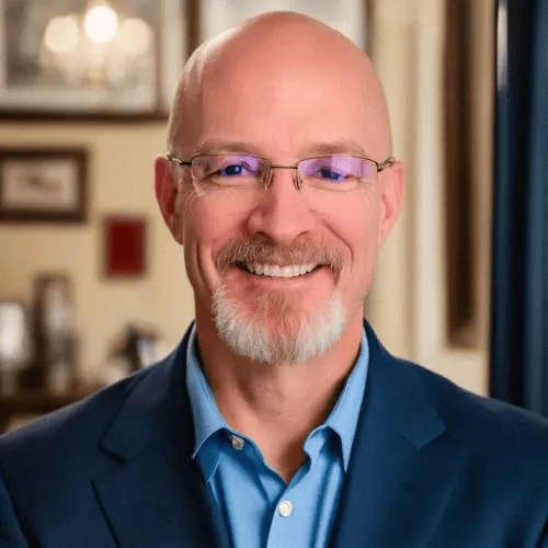 A bald man with glasses, a short white beard, and mustache, wearing a blue suit jacket and light blue shirt, smiling in a warmly lit room with blurred framed pictures in the background.
