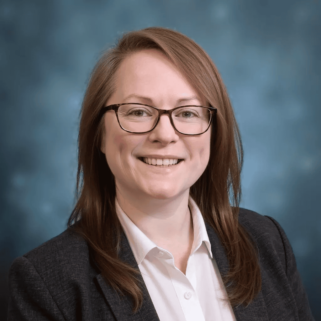 A woman with straight light brown hair and glasses smiles at the camera. She is wearing a white shirt and a dark blazer, with a blue, blurred background.